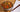 A small white bowl of fried noodles with chunks of chicken, carrots, and green onion next to a white napkin and wood chopsticks on a bamboo placemat.