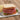 A Halal Beef salami sandwich with white bread red onion, tomato, and lettuce on a white plate with some potato chips behind to the left.