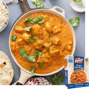 Chicken Tikka Masala with visible chunks of chicken in a ceramic pan. A bowl of rice and two pieces of naan are to the left of the pan. Cilantro is spread around the scene. the product box is overlaid in the bottom right hand corner