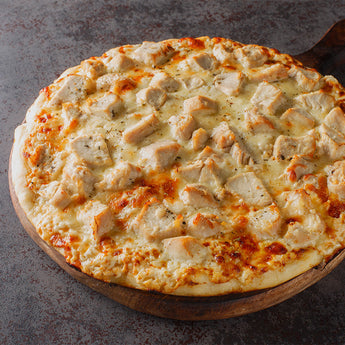 A pizza with chunks of diced chicken and cheese there some visible specs of seasoning. It is on a round wooden board.
