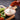 A hand dipping a piece of crispy orange-red fried chicken into a small bowl of creamy white ranch dressing, with celery sticks on a wooden board against a dark textured background.