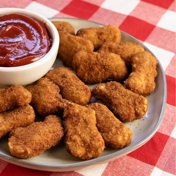 A plate of chicken nuggets stacked on a plate, a bowl of ketchup rests is on the plate to the left. A red checked cloth is under the plate.