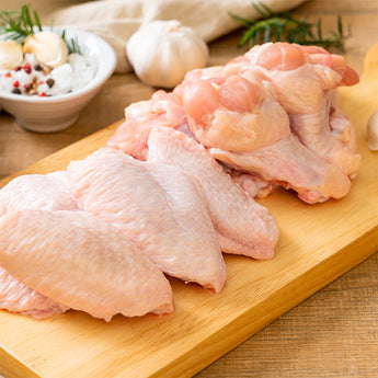 Raw chicken wings and thighs arranged on a wooden cutting board, with garlic, a small bowl of spices, and herbs in the background.