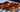 An image of an assortment of 5 skewers with chunks of red franks, green and yellow peppers, and yellow pineapple. They are served on a brown wood serving board on a blue, green, and white tablecloth in front of a black background.