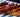 An image of an assortment of 5 skewers with chunks of red franks, green and yellow peppers, and yellow pineapple. They are served on a brown wood serving board on a blue, green, and white tablecloth in front of a black background.