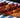 An image of an assortment of 5 skewers with chunks of red franks, green and yellow peppers, and yellow pineapple. They are served on a brown wood serving board on a blue, green, and white tablecloth in front of a black background.