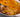 A white baking dish with a large spoon-full of cheesy potatoes, sliced thin, and mixed with pieces of crispy and savory Halal beef bacon and coated in melty cheese sauce, topped with more melted cheese. 