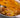 A white baking dish with a large spoon-full of cheesy potatoes, sliced thin, and mixed with pieces of crispy and savory Halal beef bacon and coated in melty cheese sauce, topped with more melted cheese. 