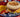 A breakfast sandwich with a slice of scrambled egg, hashbrown, and spinach, topped with melty cheese and Halal beef strips on a toasted English muffin. Served with fruit on a blue patterened plate.
