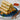 A pyramid of 10 crispy fried spring rolls sits on  plate with a small pile of sliced carrots to the left. To the right and behind the pyramid is a small square bowl of chili paste.