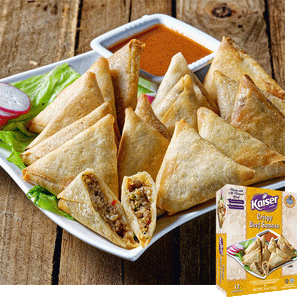 12 crispy samosas are piled together on a white plate. A samosa in front is cut open and inside is some ground beef with pieces of vegetables. A bowl of orange sauce sits behind the samosas. A box of Kaiser Crispy Beef Samosas is overtop the image. 