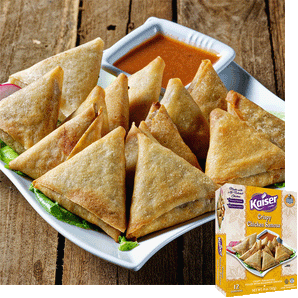 A white plate with crispy chicken samosas resting on a bed of lettuce. The samosas are garnished with a slice of radish and served with a bowl of reddish orange sauce, in the bottom right hand corner is an image of the product packaging