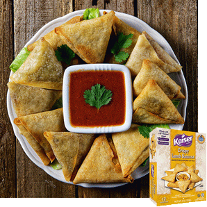 crispy lamb samosas arranged on a white plate, in the center is a square bowl with a red sauce. In the center of the sauce is a single leaf of cilantro, the rest of the plate is garnished with leaves of cilantro. The plate rests on a wooden background, in the bottom right corner is an image of the product package
