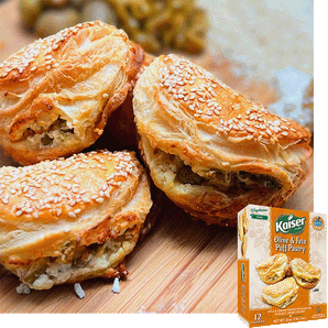 3 Kaier Olive and Feta Puff pastries are laying against each other. They have visible sesame seeds on top and feta crumbles and olive pieces coming from the opening in the flaky layers. A package of olive and feta puff pastry is overlaid in the bottom right corner. 